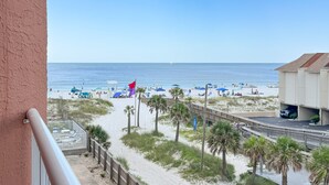A beach view from a balcony with a wooden deck, palm trees, and beach umbrellas. - Buena Vista 305 | Beachfront | Beach Side Pool in Gulf Shores | My Beach Getaways (Gulf Shores)