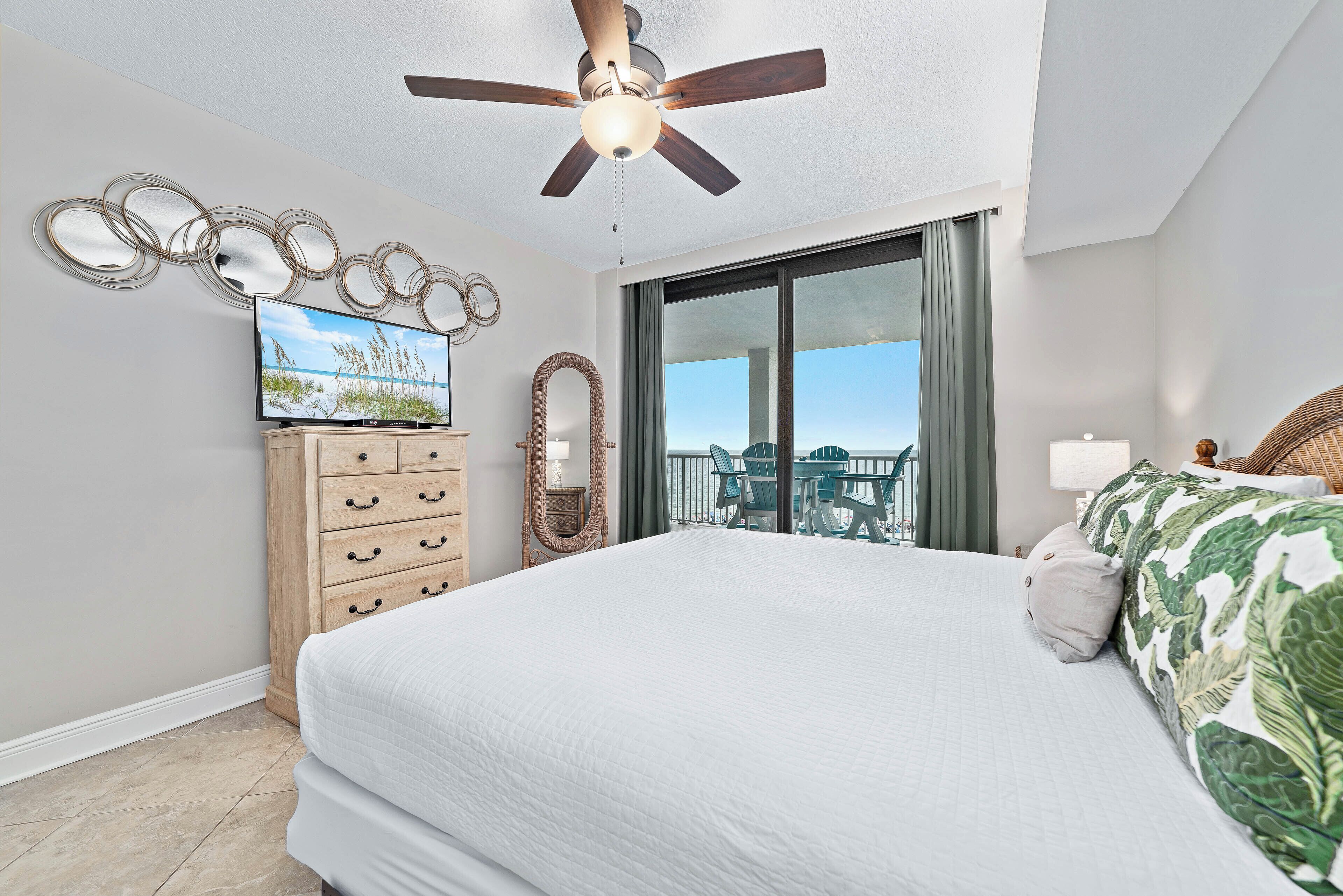 A bedroom with a large bed, a ceiling fan, a TV, a dresser, and a view of the outdoors through a sliding door.