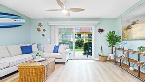 Ein modernes Wohnzimmer mit einer Couch, einem Korbtisch, einem Deckenventilator und Blick durch die Glastür nach draußen.