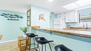 Eine Küche mit hellblauer Wand, einer Holzauslage und zwei schwarzen Barhocker. Auf der Arbeitsfläche befindet sich ein Kühlschrank, eine Spüle und ein Topf mit Pflanze.