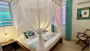 A neatly made bed with a canopy, a wooden nightstand with a lamp, a chair, and a wall-mounted picture frame. - Villa Lazy Coco, a 3-minute walk from the sea (Sainte-Luce)