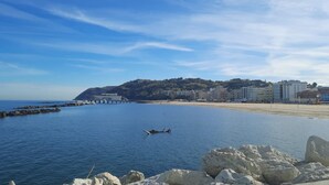 A coastal scene with a clear sky, calm sea, rocky shoreline, and buildings in the background. - GM001 - Gabicce, brand new studio apartment 30mt from the sea (Gabicce Mare)