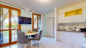 Een moderne keuken met witte kasten, een eethoek met een ronde tafel en grijze stoelen, en een groot raam met gordijnen.