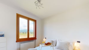 Un dormitorio con una cama grande, una mesita de noche con lámpara, una ventana que ofrece vistas al campo y un radiador debajo de la ventana.