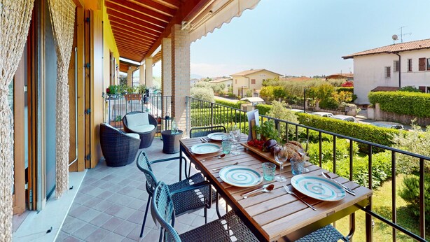 Un espacio cubierto para comer al aire libre con una mesa preparada para cuatro personas, rodeada de sillas de mimbre y vistas a un barrio residencial.