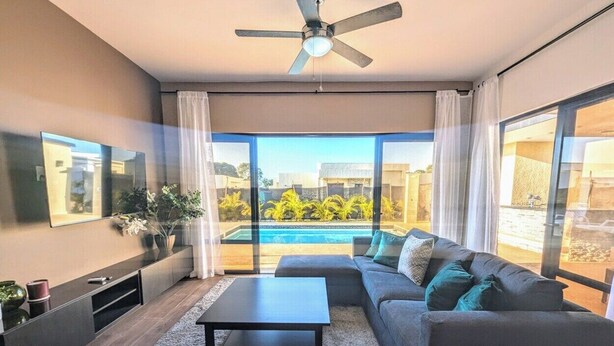 A modern living room with a sofa, coffee table, and TV. A ceiling fan is visible. The room has large windows with a view of a swimming pool and buildings. - New Sunset Villa w/Private Pool - Casa Linda (Sosúa)