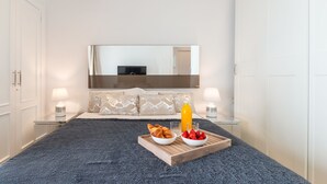 A bedroom with a large bed, a bedside table with a lamp, a tray with croissants and strawberries, and a mirror above the bed.
