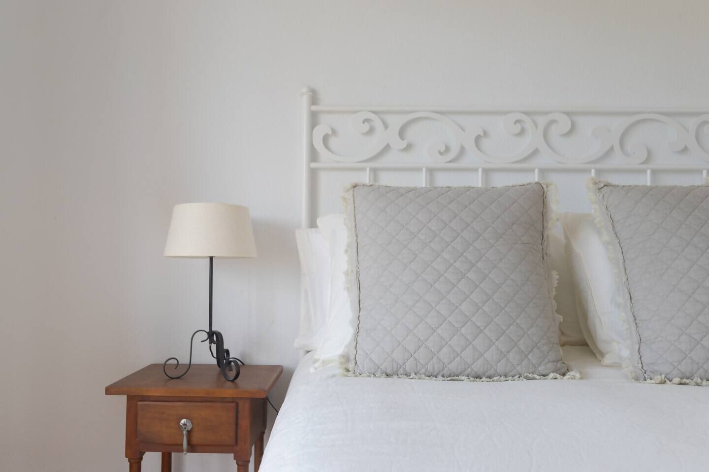 Un lit bien fait, avec une tête de lit blanche et deux oreillers, une table de chevet avec une lampe et une petite table de nuit en bois.