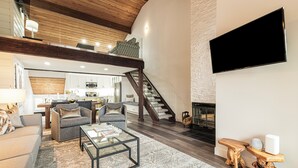 A spacious living area with a modern design, featuring a sofa, armchairs, a coffee table, and a fireplace. A staircase leads to an upper level, and a flat-screen TV is mounted on the wall.