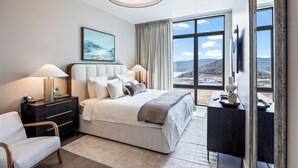 A modern hotel room with a large bed, a desk, a chair, a nightstand, and a large window offering a view of the landscape.