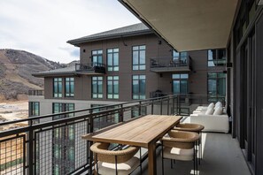Un balcón con mesa de madera y sillas de mimbre, con vista a un edificio moderno con ventanas y balcones amplios.