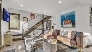 A modern living room with a sectional sofa, a coffee table, a staircase, and a large painting on the wall.