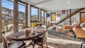 A spacious living area with a large round table, comfortable seating, a fireplace, and a view of snowy mountains through large windows.