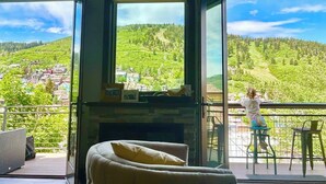A room with a fireplace, a cushioned chair, and a balcony with a view of a green hillside.