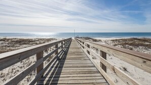 Ein hölzerner Steg führt zu einem Strand mit Dünen und blauem Himmel.