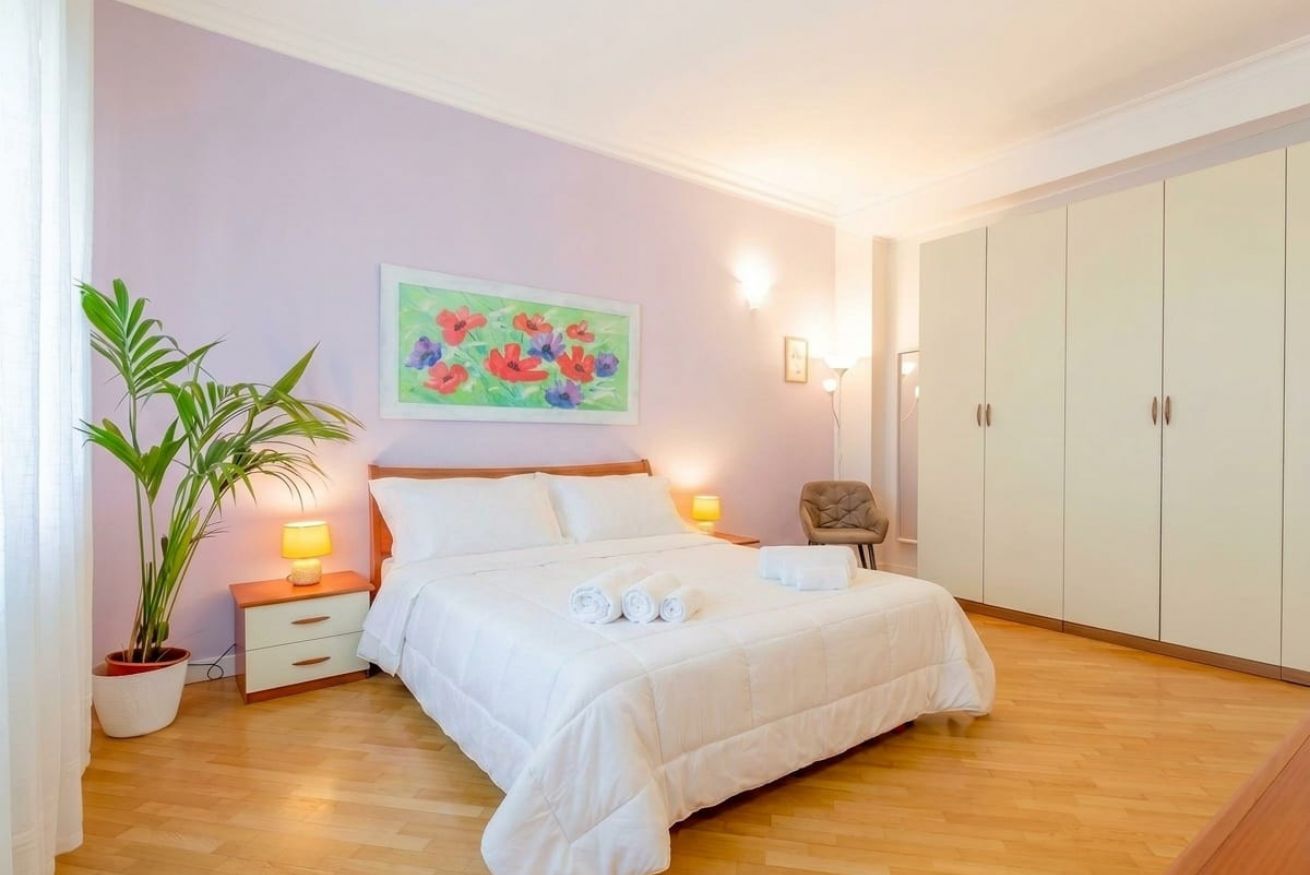 A bedroom with a bed, bedside lamps, a chair, a wardrobe, and a painting on the wall.