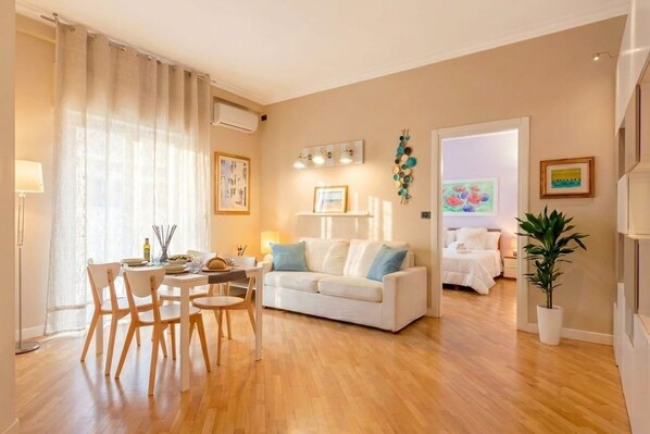Un moderno salón con un sofá blanco, mesa de comedor y sillas. Se ve una cama al fondo, una planta en maceta y obras de arte en las paredes.