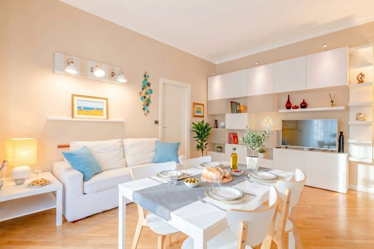 A modern living room with a dining area, white furniture, and a kitchen in the background.
