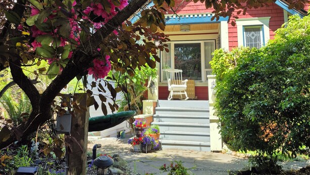 A house with a red exterior, a porch with a white chair, and a garden with pink flowers. - COZY GARDEN SUITE ON TRINITY STREET (Vancouver)