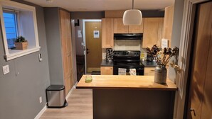 A modern kitchen with wooden cabinets, a black stove, and a wooden countertop. There is a trash can and a window with blinds. - COZY GARDEN SUITE ON TRINITY STREET (Vancouver)