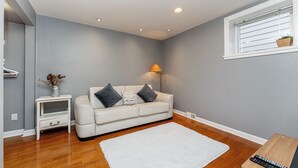 A living room with a white sofa, a small side table with a vase, a lamp, and a window with blinds. - COZY GARDEN SUITE ON TRINITY STREET (Vancouver)