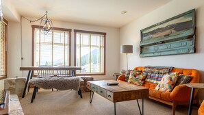 A cozy living room with a wooden coffee table, a colorful sofa, and a large window with blinds. - Ski In/Out with Mountainside Views. Dog Friendly (Fernie)