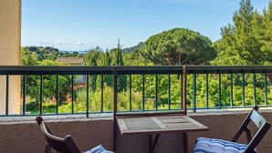 Un balcon avec une table et deux chaises, donnant sur un paysage verdoyant, avec des arbres et une vue lointaine sur des bâtiments.