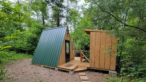 Una pequeña cabaña de madera con techo verde y porche de madera, rodeada de un bosque denso.