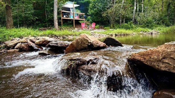 Una cabaña rústica con un balcón de madera y un pequeño porche, rodeada de un bosque y un arroyo.