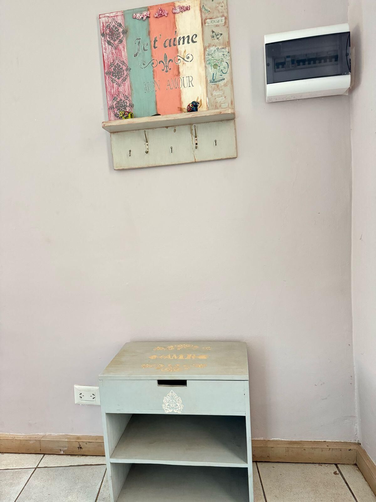 Un piccolo tavolo di legno blu chiaro con un cassetto, un elemento decorativo appeso al muro sopra e una scatola elettrica bianca sulla parete.