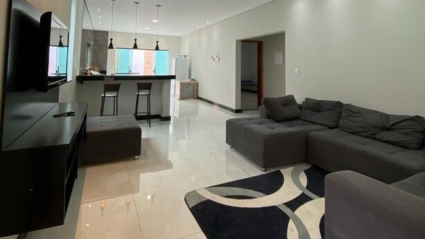 A modern living room with a large sectional sofa, a flat-screen TV mounted on the wall, a kitchen area with bar stools, and recessed ceiling lighting. - Complete House for Rest (Pedro Leopoldo)