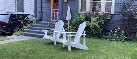Et hus med blå facade, en veranda med trin og to hvide Adirondack-stole på plænen.