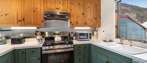 Een moderne keuken met houten kastjes, roestvrijstalen apparatuur en een groot raam met uitzicht op een huis en heuvels.
