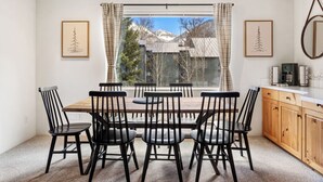 A dining area with a wooden table and black chairs, a window with curtains, and a kitchen with wooden cabinets and a coffee machine. - Experience luxury at Alpenglow at Telluride Lodge, a spacious 2 bed/2-bath condo (Telluride)