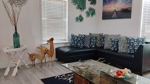 Een moderne woonkamer met een zwarte bank, decoratieve kussens, een glazen salontafel en een schilderij van een zonsondergang.