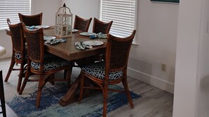 Een eetruimte met een houten tafel, rieten stoelen en een blauw vloerkleed. Er zijn twee ramen met jaloezieën, een plafondlamp en wanddecoratie.