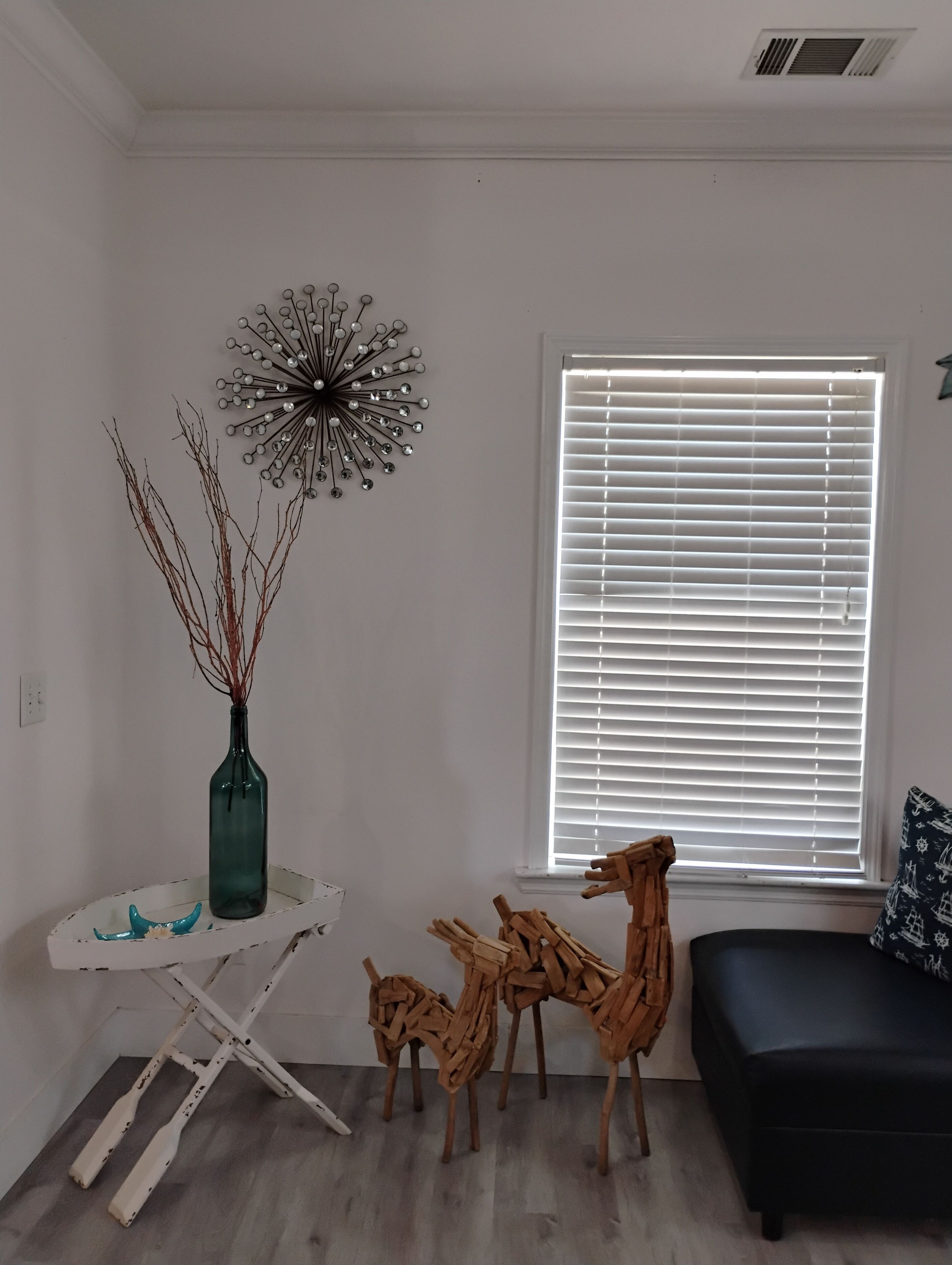 Een woonkamer met een zwarte bank, een houten sculptuur, een decoratief metalen object en een raam met zonwering.