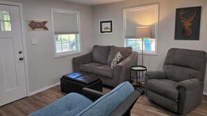 Un salon avec un canapé gris, un banc bleu et un fauteuil gris. Il y a deux fenêtres avec des stores, une lampe et une œuvre d’art représentant une tête de cerf accrochée au mur.