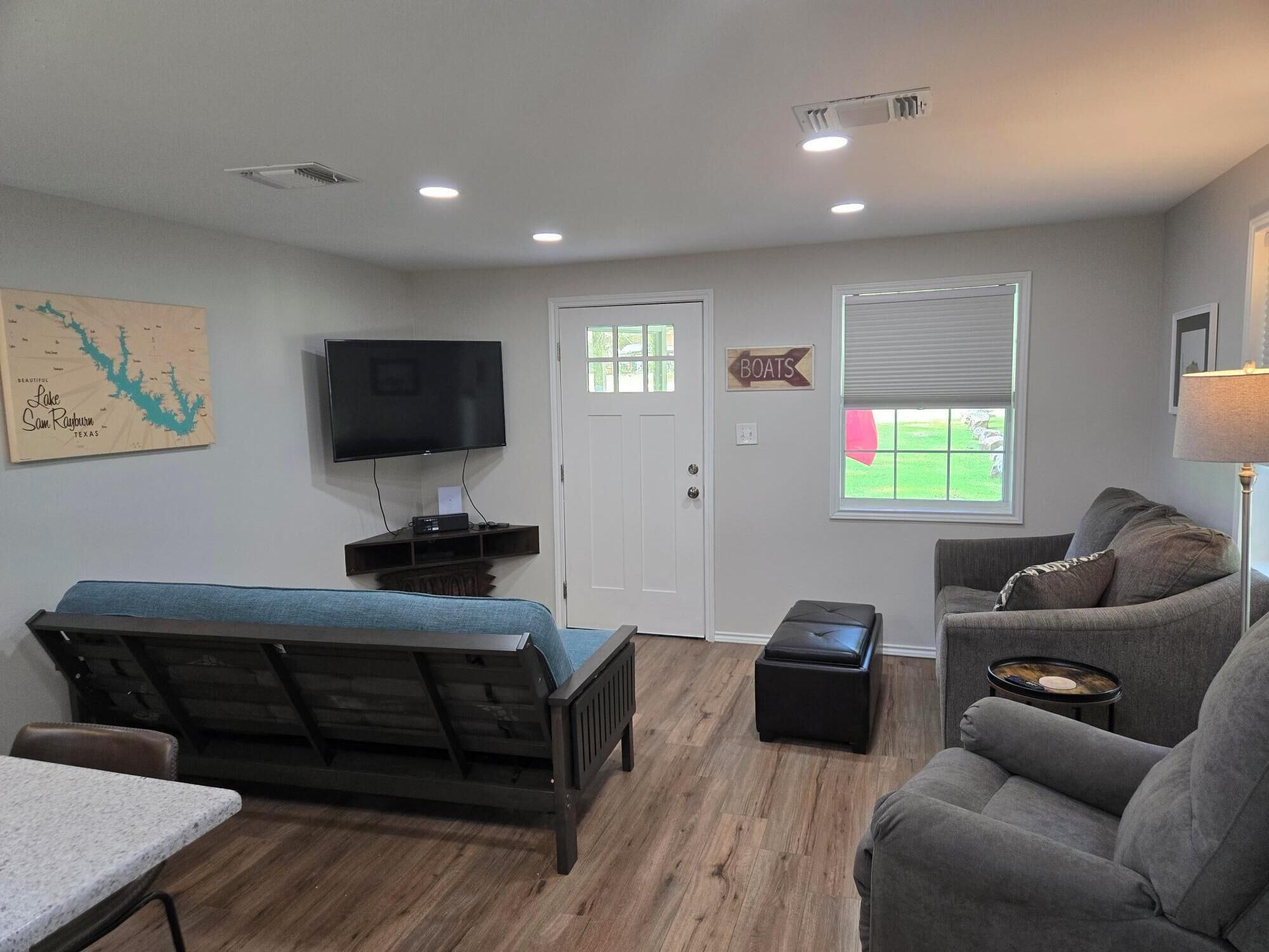 A living room with a sofa, a TV mounted on the wall, a wooden bench, a small table, and a window with blinds.