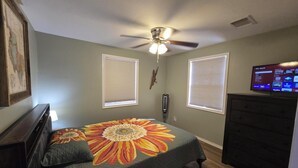 Une chambre à coucher comprenant un lit, une commode, un ventilateur de plafond, deux fenêtres équipées de stores et un téléviseur posé sur la commode.