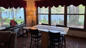 Ein Zimmer mit einem Klavier, einem Schreibtisch und zwei Stühlen. Große Fenster mit Vorhängen und eine Lampe auf dem Schreibtisch.
