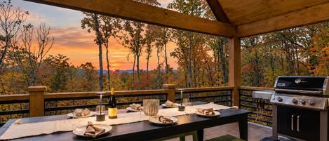 An outdoor dining area with a view of trees and a sunset, a grill, and a table set for two.