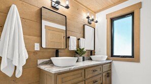 A bathroom with wooden panel walls, a large mirror, two white vessel sinks, and a window with a view.