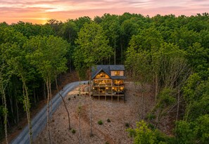 A two-story cabin with a deck, surrounded by dense forest, at sunset.