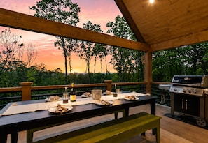 An outdoor dining area with a table set for two, a grill, and a view of trees and a sunset.