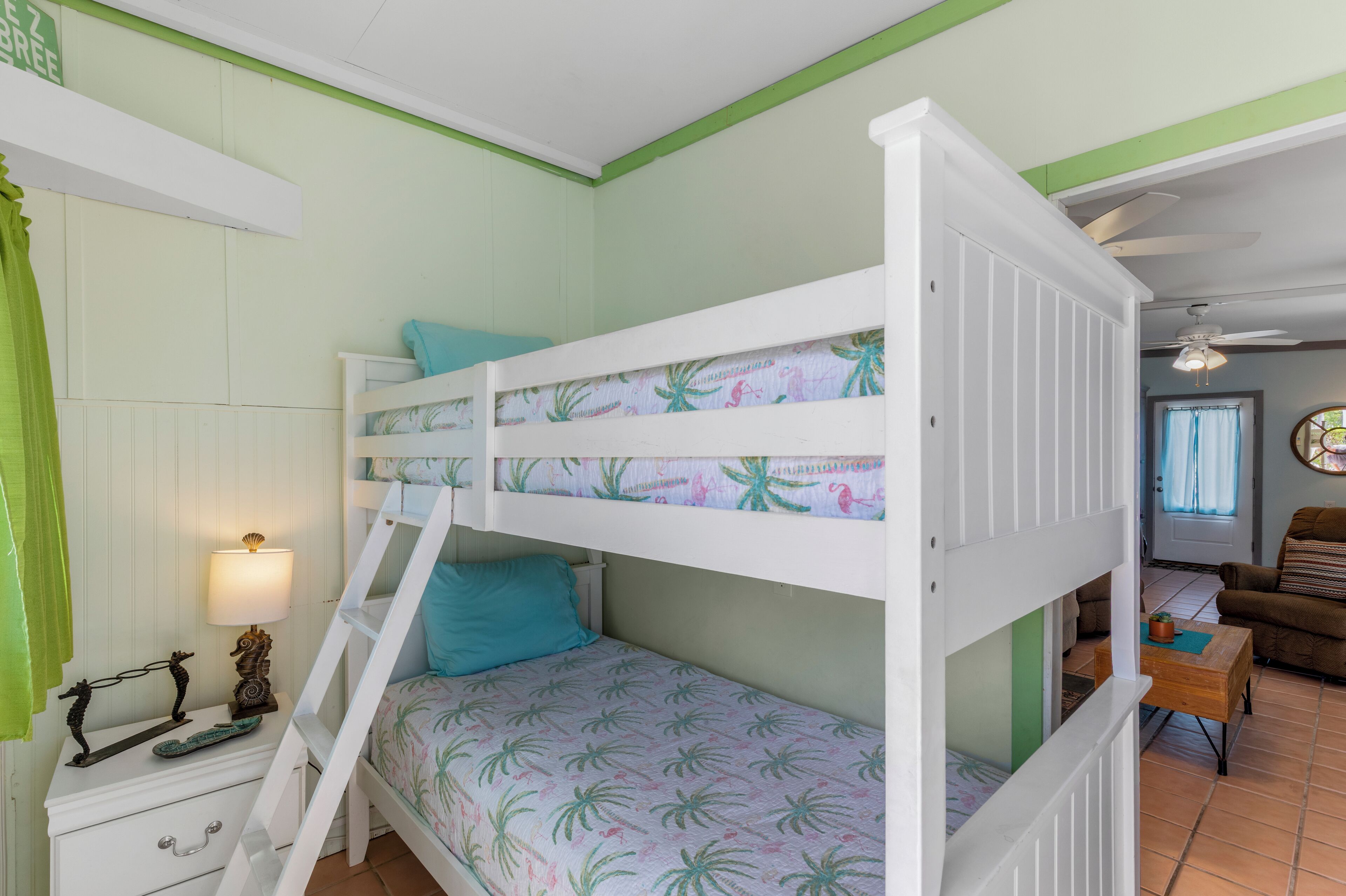 Una cama litera con cubrecama de temática tropical, una mesita de noche con lámpara y una cortina verde.