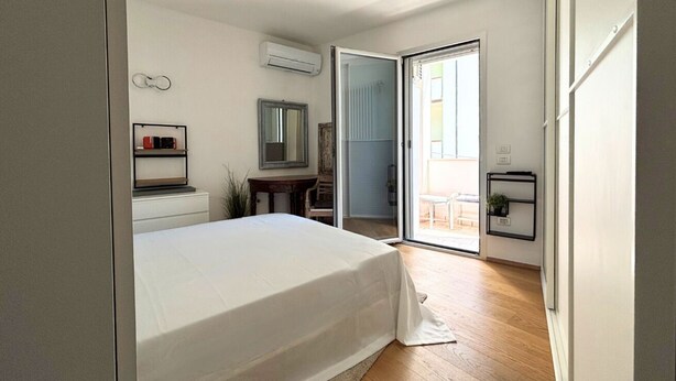 Un dormitorio con cama, cómoda, espejo, equipo de aire acondicionado, puerta corrediza de vidrio que da a un balcón y una mesita con una planta.