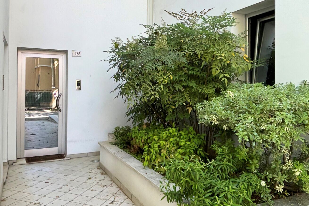 Une entrée d’immeuble avec une porte portant le numéro « 25 », un sol carrelé et un petit espace jardin agrémenté de différentes plantes.