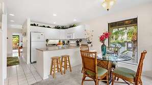 A modern kitchen with white cabinets, a central island, wooden stools, and a dining table with chairs. - Cozy ground floor condo, lush lanai, dining nearby (Kihei)