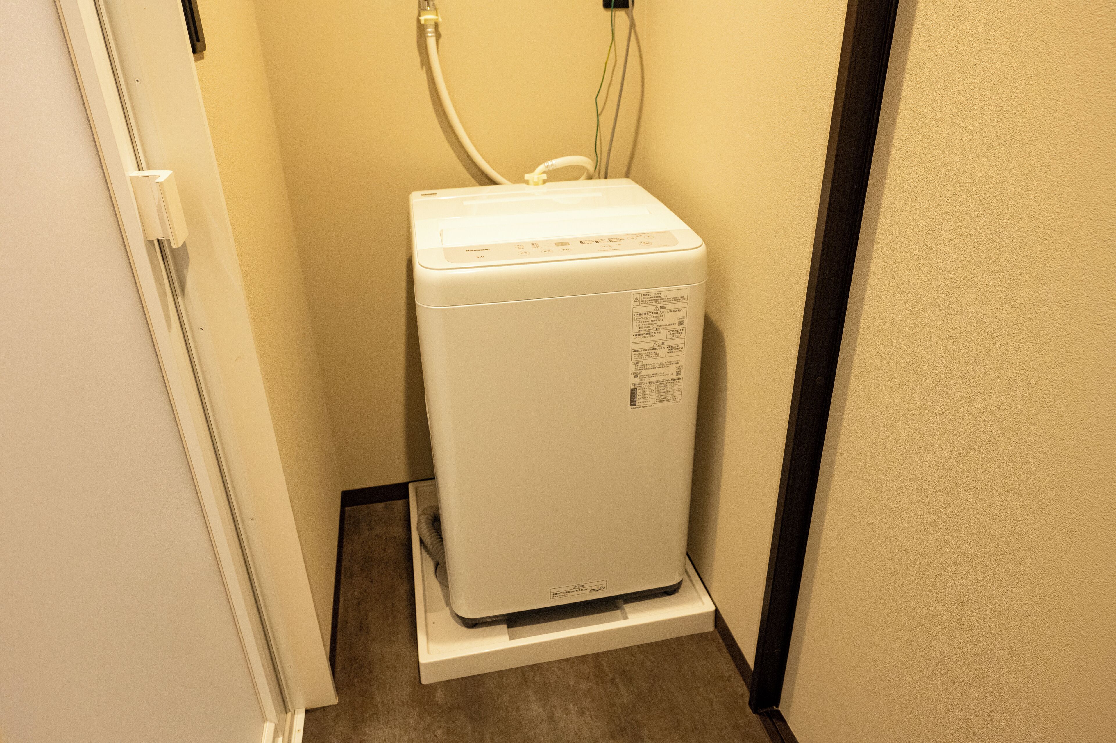 A compact, white, wall-mounted washing machine in a narrow hallway with a door on one side and a dark floor.